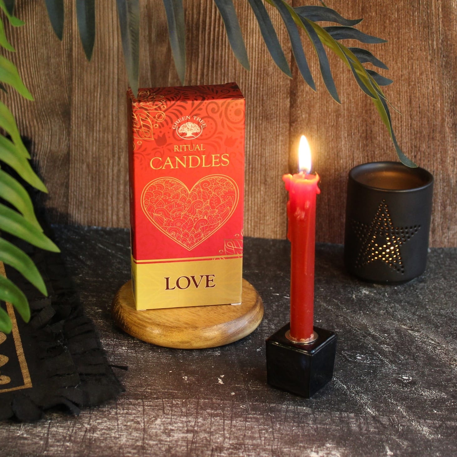 A boxed Ritual Candles product labeled 'LOVE' stands on a small wooden base beside a lit red candle in a black holder, with a dark star-shaped candle holder and green leaves against a rustic wood background.