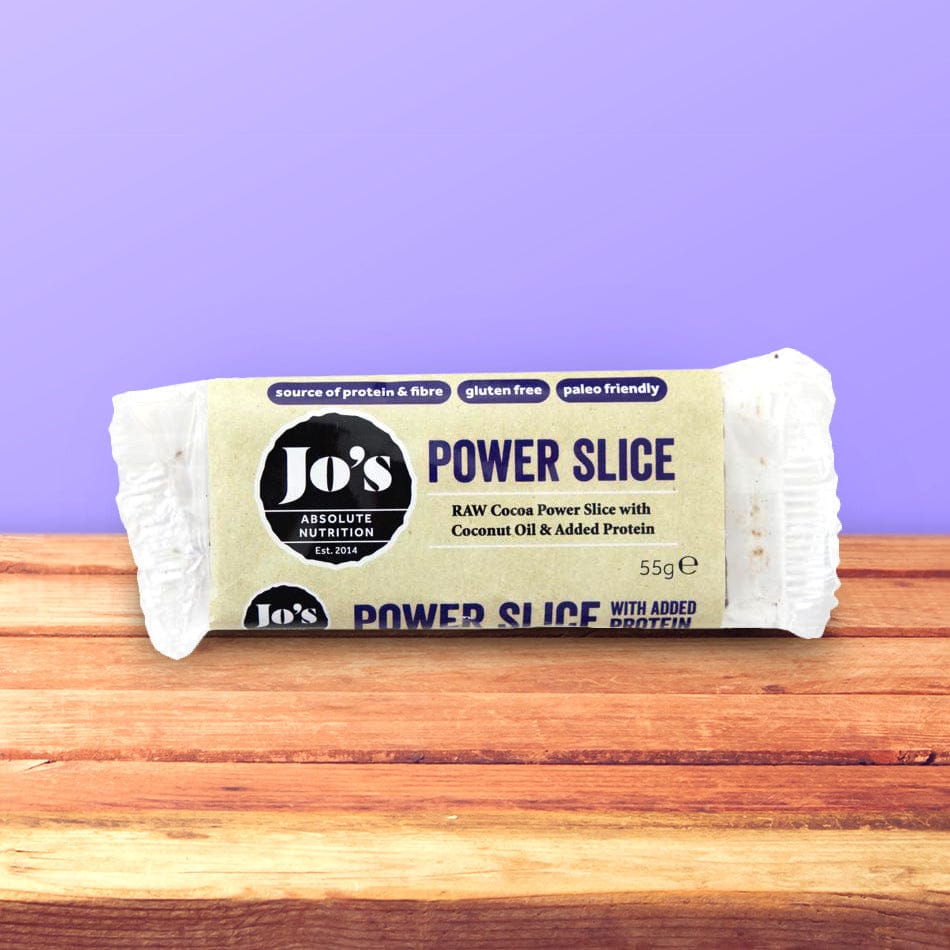 A Jo's Power Slice protein bar wrapped in a kraft paper sleeve, resting on a wooden surface against a solid purple background.