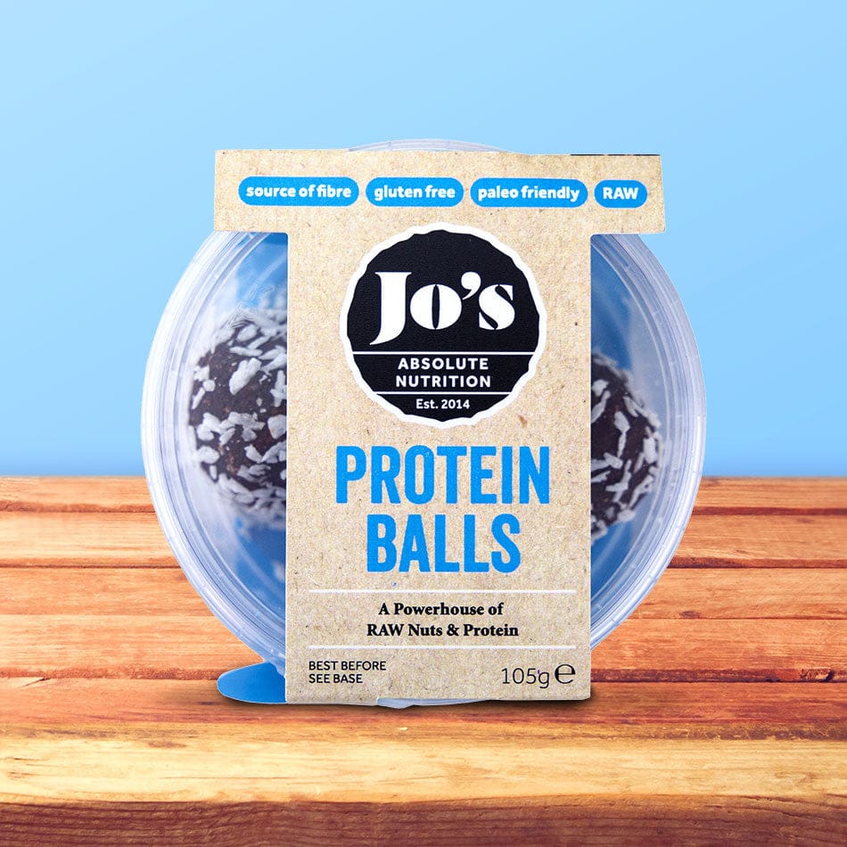 Jo's Absolute Nutrition Protein Balls in a clear plastic tub with a kraft paper label, displayed on a wooden surface against a light blue background.