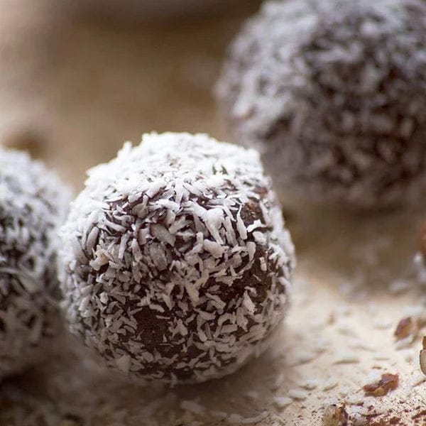 Close-up of chocolate coconut balls coated in white shredded coconut, with one ball in sharp focus in the foreground and two blurred balls in the background on a light surface.