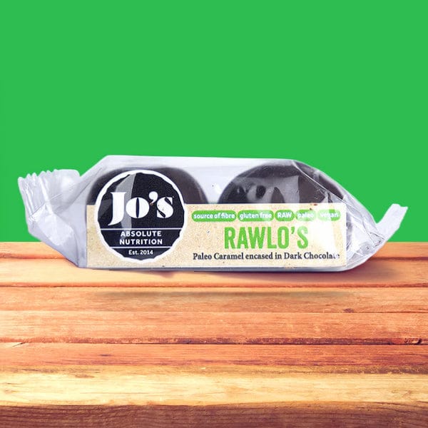 A packaged food product labeled Jo's Rawlo's Paleo Caramel Encafed in Dark Chocolates, shown in clear wrapping on a wooden surface against a bright green background.