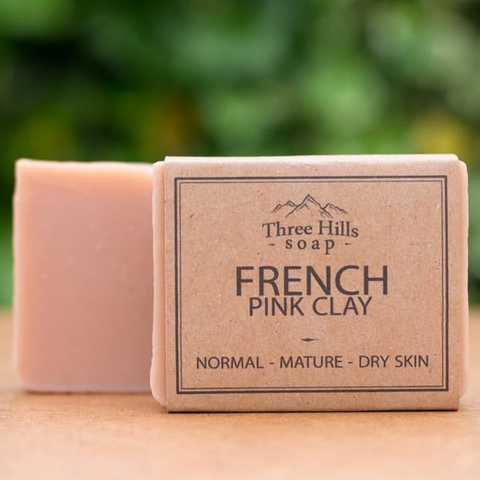 A bar of French Pink Clay soap by Three Hills Soap, shown in its kraft paper packaging box with the bar partially visible behind it, set against a blurred green background.