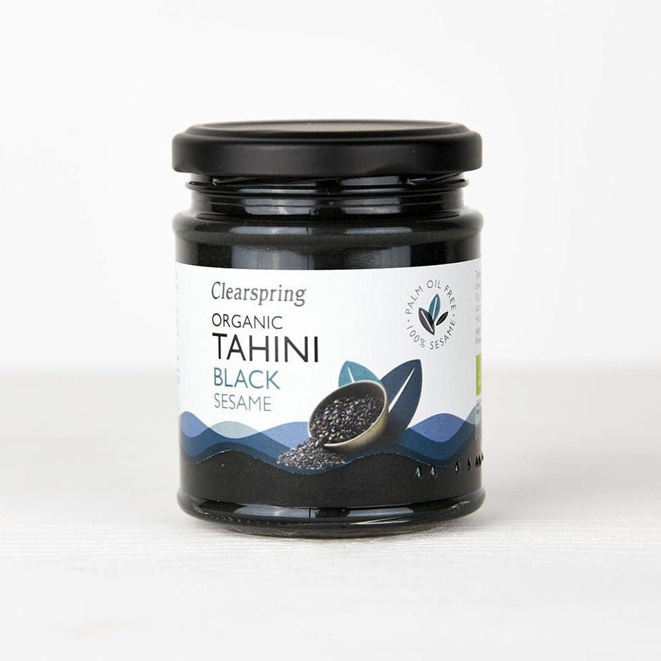 A jar of Clearspring Organic Tahini Black Sesame on a white background. The product is in a clear glass jar showing the dark black sesame paste inside, with a black screw-on lid and a white label featuring blue and black graphics.