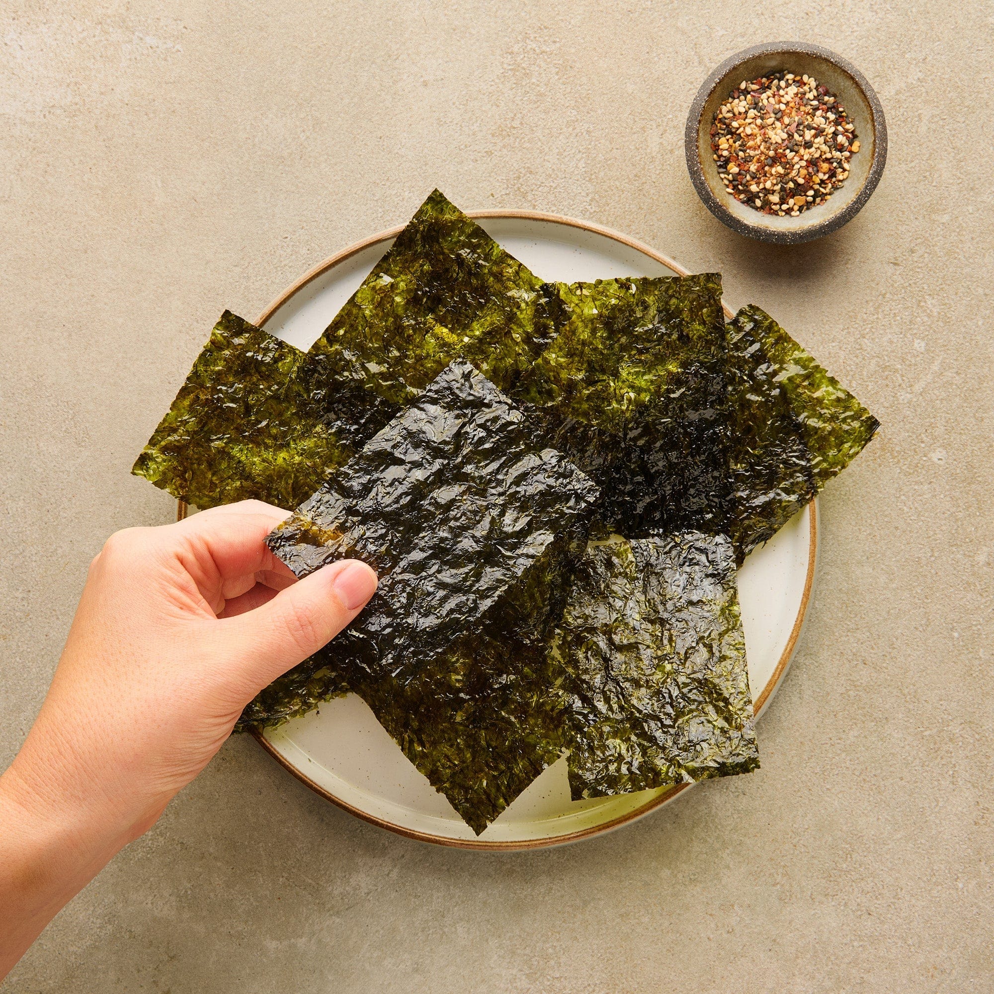 Hand holding a piece of seaweed over a plate with more seaweed and a small bowl of seasoning on a beige surface.