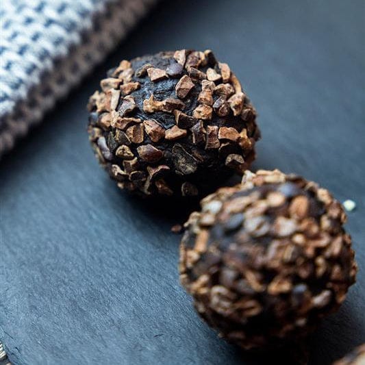 Two chocolate-covered truffles coated in chopped dark chocolate and nuts, arranged on a dark slate surface with a textured cloth in the background.