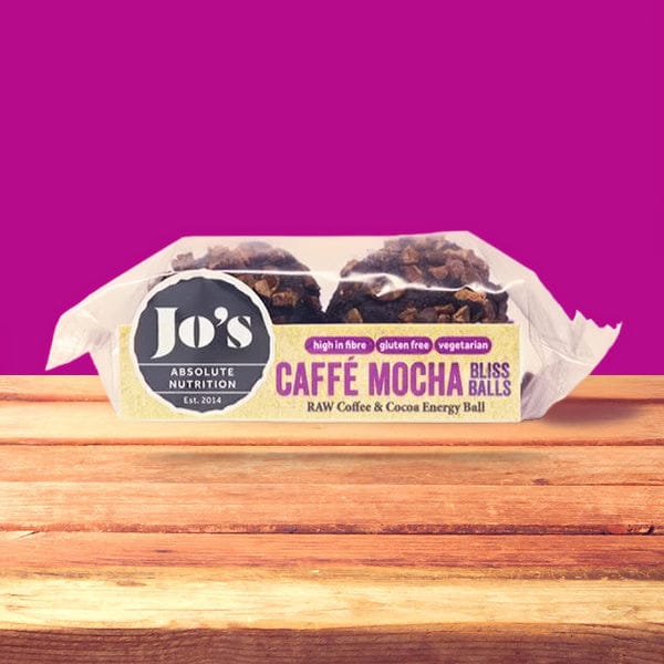 A packaged Jo's Absolute Nutrition Caffe Mocha Bliss Ball energy ball on a wooden surface against a bright magenta background.