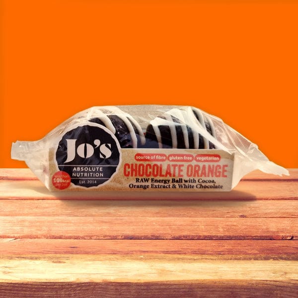 A packaged Jo's Absolute Nutrition Chocolate Orange raw energy ball, wrapped in clear plastic with a printed label, resting on a wooden surface against a solid orange background.