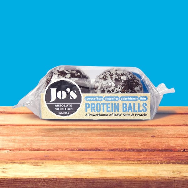 A package of Jo's Absolute Nutrition Protein Balls placed on a wooden surface against a bright blue background. The clear plastic wrapper shows two dark chocolate protein balls coated with white flakes, with a label reading 'Jo's Protein Balls'.