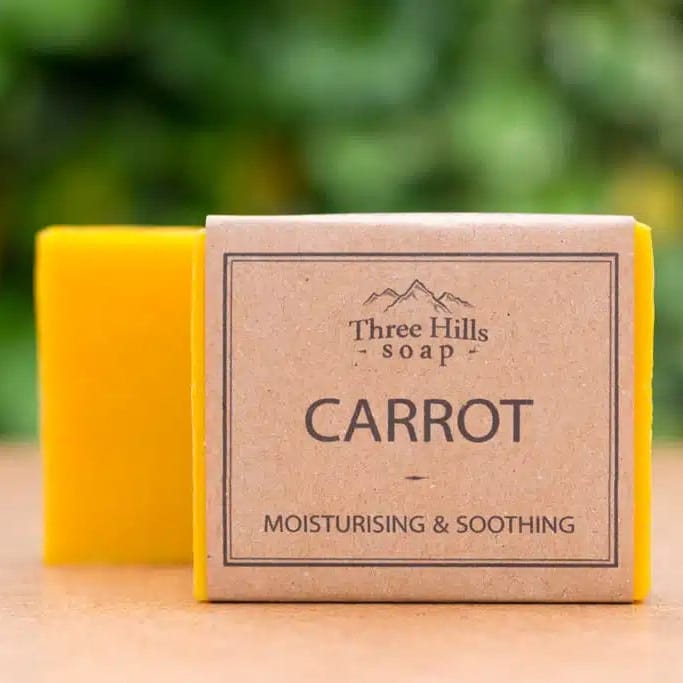 A bar of Three Hills Soap in the Carrot scent, wrapped in a brown kraft paper sleeve, with a bright yellow soap bar visible behind it on a wooden surface with a blurred green background.