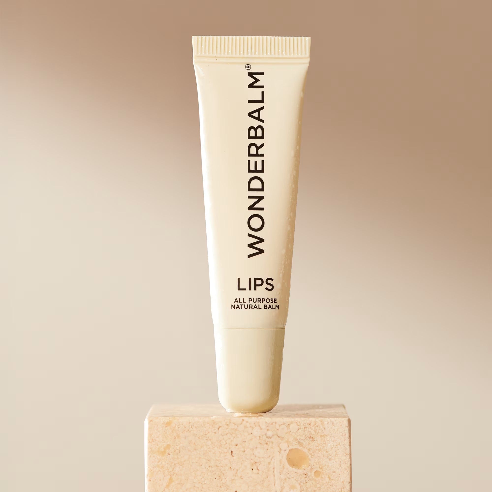 A cream-colored tube of Wonderbalm lip balm stands upright on a small stone pedestal against a warm beige gradient background. The tube features black text reading 'WONDERBALM LIPS ALL PURPOSE NATURAL BALM'.